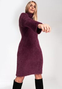 Plüschiges, strukturiertes lila Kleid mit hohem Kragen und langen, ausgestellten Ärmeln. Der Saum ist über dem Knie, kombiniert mit schwarzen kniehöhen Stiefeln.