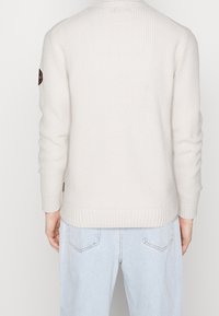 Vaaleanbeige neulepaita, jossa on resorikuvio, pitkät hihat ja pyöreä pääntie. Vasemmassa olkapäässä on logoplässi.