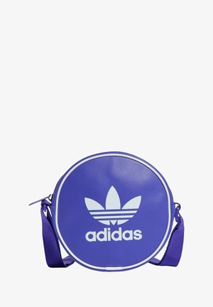 Okrągła fioletowa torba Adidas z białym logo trefoil i regulowanym paskiem na ramię na białym tle.
