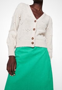 Cardigan a maglia color crema con scollo a V, dettagli a pallini testurizzati e bottoni in legno, abbinato a una gonna in velluto a coste verde brillante.