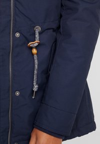 Mörkblå vadderad jacka med dragkedja, som har en texturerad ärm, flätad dragsko med trädetaljer och tryckknappar.