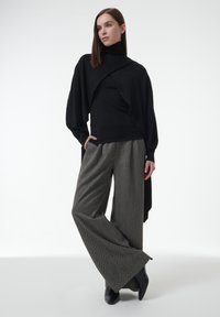 Schwarzer Rollkragenpullover mit drapiertem Vorderteil, kombiniert mit weit geschnittenen, strukturierten grauen Hosen. Schwarze Knöchelstiefel runden das Outfit ab. Minimalistisches Design.