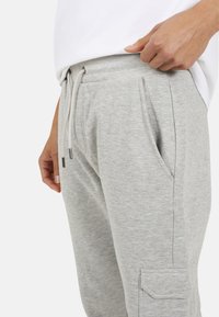 Jasnoszare spodnie dresowe z elastycznym pasem, sznurkiem, bocznymi kieszeniami oraz kieszenią cargo na lewej nodze. Miękki, teksturowany materiał.