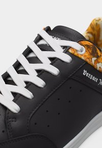 Μαύρο δερμάτινο αθλητικό παπούτσι με λευκά κορδόνια, διάτρητη πλευρά, λογότυπο "Versace Jeans" και πορτοκαλί υφασμάτινο σχέδιο γύρω από το κολάρο του αστραγάλου.