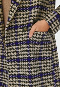 Hand met ongebroken nagels in de zijzak van een houndstooth-patronen jas in zwart, beige en paars gestoken.