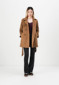 Manteau en suède marron avec un design croisé, deux poches avant et une ceinture à la taille. Le modèle porte un débardeur noir et un jean évasé noir.