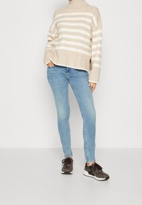 Osoba w beżowym i białym swetrze w paski z golfem, jasnoniebieskich obcisłych dżinsach i ciemno brązowych trampkach, stojąca z jedną ręką w kieszeni.