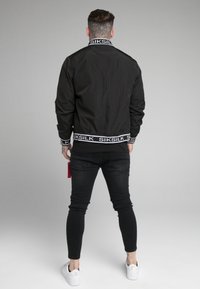 Mu muž s tetováním na krku, oblečený v černé bundičce Siksilk, černých skinny džínech a bílých teniskách, stojí s zády otočenými k šedému pozadí.