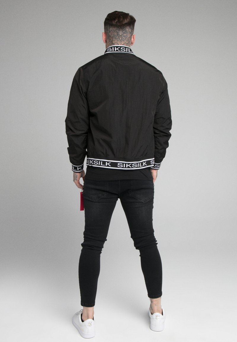 Mu muž s tetováním na krku, oblečený v černé bundičce Siksilk, černých skinny džínech a bílých teniskách, stojí s zády otočenými k šedému pozadí.