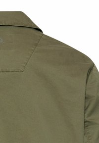 Olijfgroene jas, achterkant toont kraag en schoudernaad met subtiele gestikte details op de stof.