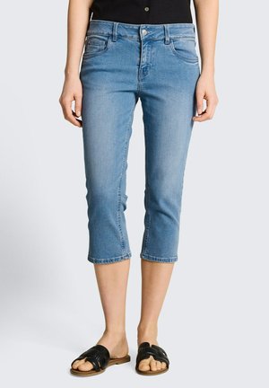 Vrouw draagt lichtblauwe cropped spijkerbroek en zwarte slides, staand tegen een effen achtergrond.