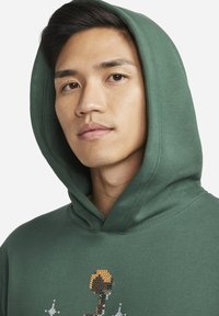 Felpa verde con un grande cappuccio, caratterizzata da un design grafico pixelato in arancione, marrone e bianco sulla parte anteriore. Tessuto a trama liscia.