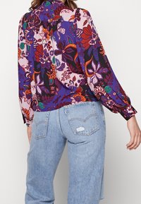 Donna che indossa jeans in denim blu chiaro e una blouse floreale a maniche lunghe con motivi viola, rossi, arancioni e verdi, vista da dietro.