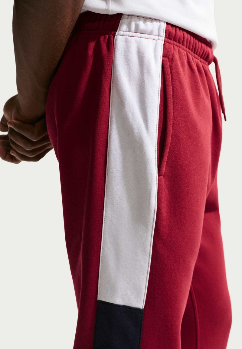 Vista laterale di una persona che indossa pantaloni della tuta rossi con una striscia verticale bianca e blu navy, mani giunte dietro la schiena.