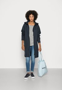 Chaqueta larga azul marino con capucha, camiseta gris debajo, bolsa tote azul claro, vaqueros azules y zapatillas oscuras con suelas blancas. Textura de tela suave.