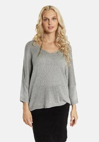 Pilkas megztinis su V formos iškirpte, trijų ketvirčių rankovėmis ir laisvu siluetu, pasižymintis blizgia tekstūra ir giriamais kraštais. Užsidėtas ant juodos sijono.