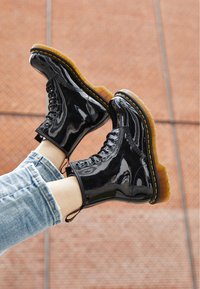 Botines de charol negro con costuras amarillas, diseño con cordones y suela de goma translúcida. Combinados con jeans azul claro.