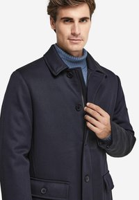 Mörkblå ullblandad kappa med skräddarsydd design, med slagkrage, två framfickor och knäppning med knappar. Lager på en blå polotröja.