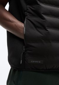 Το χέρι ενός ατόμου έχει εισαχθεί στην πλευρική τσέπη ενός μαύρου επενδεδυμένου μπουφάν με εμφανές φερμουάρ και το έμβλημα με το λογότυπο Icepeak.