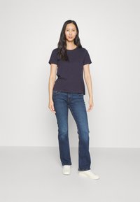 Granatowa koszulka z okrągłym dekoltem, bawełniana, zestawiona z niebieskimi dzwonami. Model nosi białe trampki, stojąc na tle jasnoszarym.