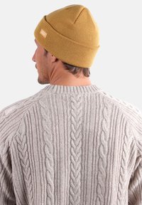 Bonnet en tricot jaune moutarde au design sans couture avec une petite étiquette logo, associé à un pull en tricot gris avec des motifs en relief.