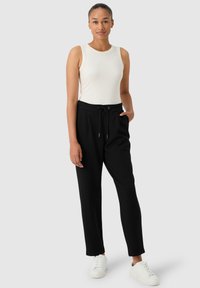 Pantalones joggers negros ajustados con cintura con cordón, combinados con una blusa sin mangas en tonos neutros. El modelo lleva zapatillas blancas y está de pie frente a un fondo sencillo.