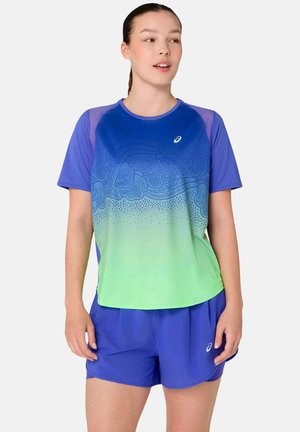 Frau trägt ein blau-grün verlaufendes Sportshirt mit abstrakten Mustern und passende blaue Shorts, steht vor einem einfarbigen Hintergrund.