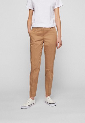 Osoba ubrana w beżowe, dopasowane spodnie, białą koszulkę z okrągłym dekoltem oraz białe tenisówki z płótna, na jednolitym tle.