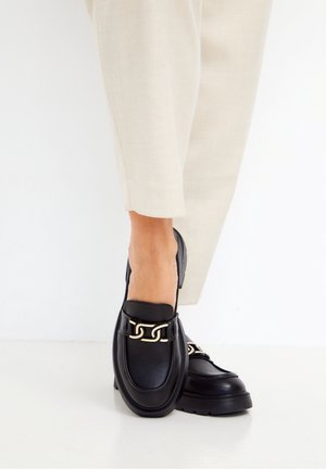 Person trägt schwarze Leder-Loafers mit goldener Kettendetail und cremefarbene Hose, steht auf einem glatten weißen Boden.