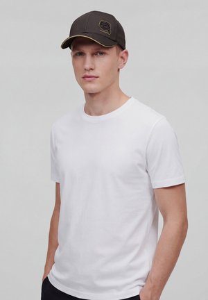 Man draagt een wit T-shirt met korte mouwen en een donkere honkbalpet, staat met handen in de zakken, neutrale grijze achtergrond.