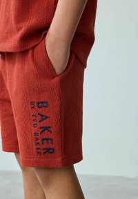 Červené texturované šortky s bočními kapsami a modrou vyšívanou nášivkou "BAKER by Ted Baker" na levé noze. Čistý, pohodlný design.