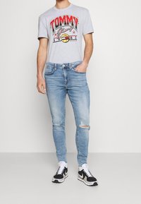 Mies, joka käyttää vaaleansinisiä revittyjä slim fit -farkkuja, mustia ja valkoisia lenkkikenkiä sekä harmaata Tommy Jeans -t-paitaa, jossa on punaisia ja keltaisia kuvastoja.