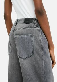 Person wearing high-waisted, loose-fit washed gray denim jeans with large back pockets and a white tucked-in ribbed top.