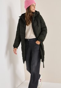 Cappotto lungo imbottito nero con cappuccio, abbinato a un maglione crema, pantaloni grigio scuro e un berretto lavorato a maglia rosa. Presenta una chiusura con zip.