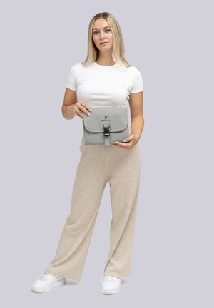 Femme en t-shirt blanc et pantalon beige tenant un sac pliable gris avec fermoir devant un fond uni.