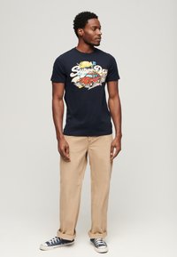 Camiseta navy con estampado gráfico que presenta un coche rojo y texto, combinada con pantalones beige claros y zapatillas azules. Manga corta, cuello redondo.