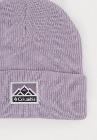 Violeta adīta cepure ar locītavu un taisnstūra Columbia logotipa uzlīmi, kurā attēloti kalnu ikonas melnā un baltā krāsā.