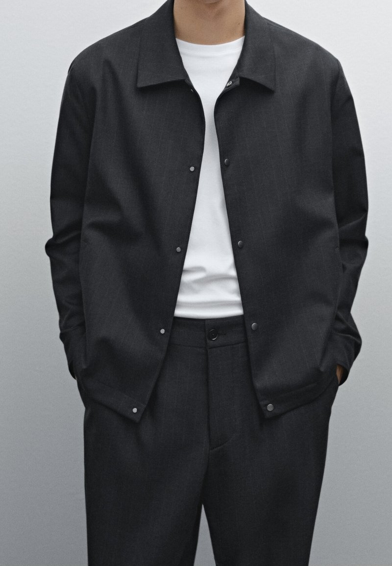 Personne portant une veste boutonnée gris foncé sur un t-shirt blanc et un pantalon gris foncé assorti, mains dans les poches, fond neutre.