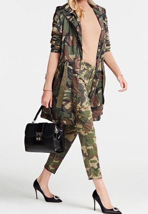 Femme portant une veste et un pantalon camouflage, un haut beige à manches longues, des escarpins noirs à boucle, tenant un sac à main noir avec un logo.
