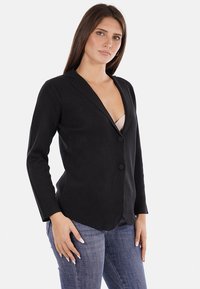Blazer nero con un design aderente, chiusura a singolo bottone, reverse a lancia e maniche lunghe, abbinato a jeans in denim. Tessuto di consistenza liscia.