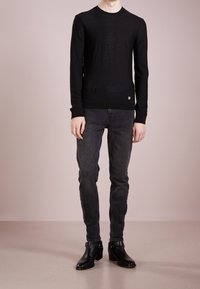 Svart lätt stickad tröja med långa ärmar, rund halsringning och subtil logotypdetalj vid fållen, i kombination med mörkgrå tajta jeans och svarta stövlar.