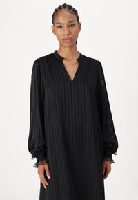 Črna obleka z dolgimi rokavi in vertikalnimi črtami, z V-izrezom in prozornimi napihnjenimi rokavi. Okrašena z čipkastim obrobom na manšetah.