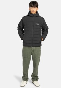 Giacca a vento nera con cappuccio e design a cuciture orizzontali, dotata di zip frontale e patch con logo. Abbinata a pantaloni cargo verdi.