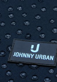 Waterdruppels op een gestructureerde donkere stof met een rechthoekige patch met het label "JOHNNY URBAN" en een gestileerd "J"-logo.