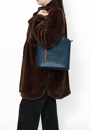 Borsa a secchiello in pelle blu scuro con texture perforata, dotata di chiusura zip e tracolla color cuoio. Indossata sopra un cappotto marrone.