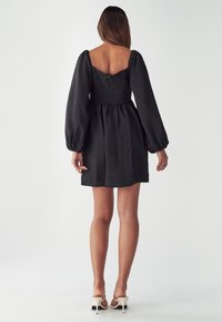 Schwarz Kleid mit einem strukturierten Stoff, Puffärmeln und einer gerafften Taille. Es hat einen quadratischen Rückenausschnitt und einen Reißverschluss.