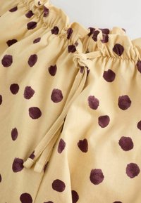 Gelber Stoff mit unregelmäßigen dunkelvioletten Tupfen, ausgestattet mit einem gerafften elastischen Bund und einem gebundenen Kordelverschluss.