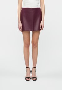 Vrouw in een donkerpaarse leren mini-jurk, witte top en zwarte enkelband hoge hakken, staand tegen een effen witte achtergrond.