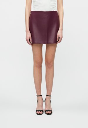 Woman wearing a dark purple leather mini skirt, white top, and black ankle-strap high heel sandals, standing against a plain white background.