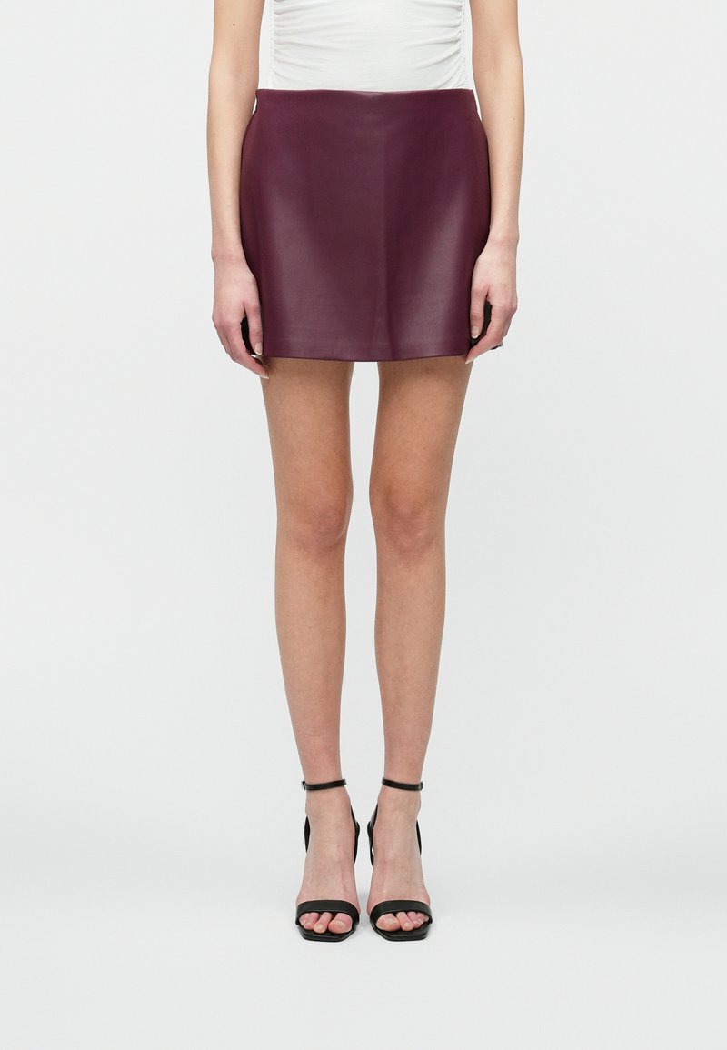 Vrouw in een donkerpaarse leren mini-jurk, witte top en zwarte enkelband hoge hakken, staand tegen een effen witte achtergrond.